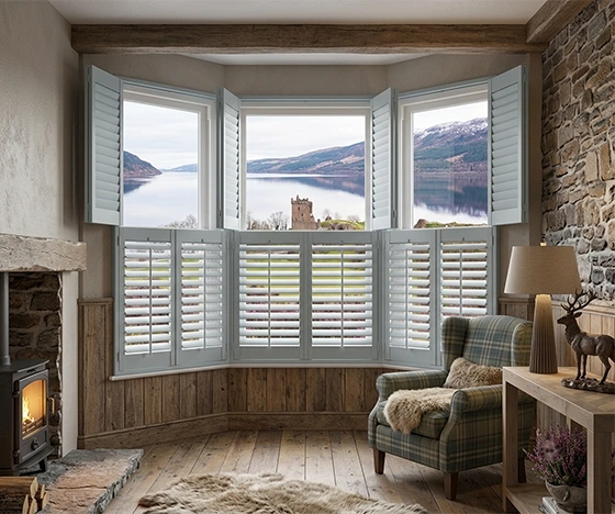 Tier on Tier shutters, specific style shutter kensington skyline grey tier main standard.webp, installed on a large three-window bay in a traditional Highland living room with stone and wood details. The lower shutters are closed, and the upper shutters are swung wide open to frame a panoramic view of a loch, Urquhart Castle ruins, and mountains under a natural daylight sky