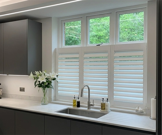 A modern grey handleless kitchen featuring a large window with white cafe style plantation shutters installed. A vase of white lilies and a black stand mixer sit on the light quartz countertop next to an undermount sink with a sophisticated faucet.